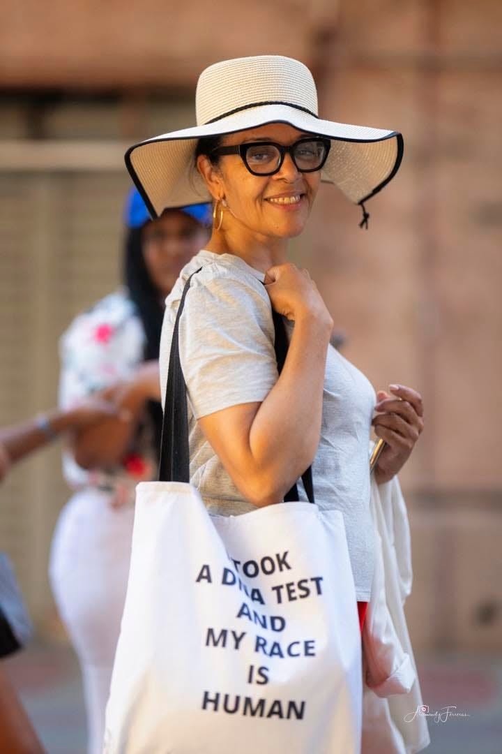 Fe Bencosme wearing a wide-brimmed hat and carrying a tote that reads, I took a DNA test and my race is human